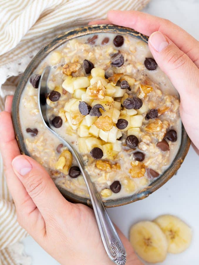 Hands holding a bowl of Overnight Oats topped with banana, nuts and chocolate