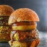 A big, thick Down Home Vegan BBQ burger in a seeded bun with an onion ring, slice of tomato and lots of BBQ sauce.