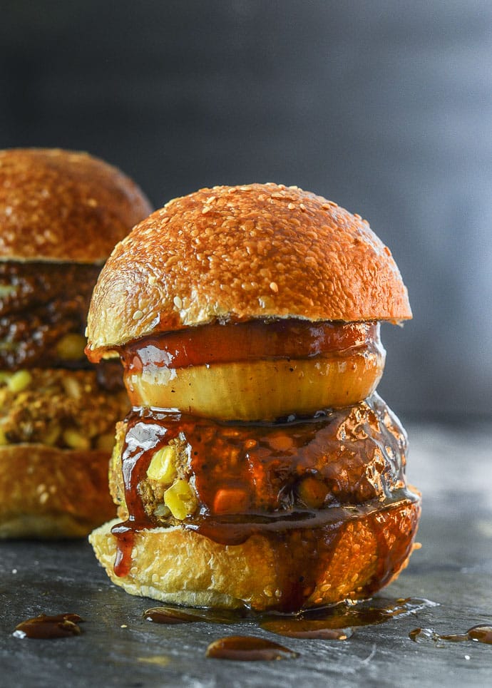 A big, thick Down Home Vegan BBQ burger in a seeded bun with an onion ring, slice of tomato and lots of BBQ sauce.